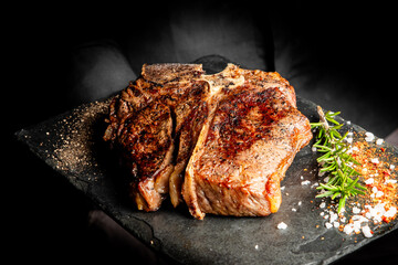 Grilled T-bone steak seasoned with coarse salt, cracked pepper, and fresh rosemary on a black slate board, dark background, close-up, food photography