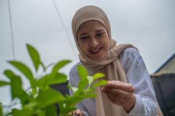 Asian muslim woman leans to inspect a vibrant shrub in a sunlit garden, embodying gardening.