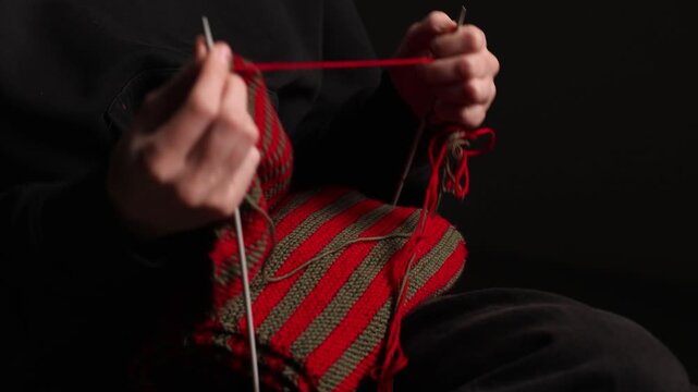 Close-up of a person knitting a red and green striped fabric with needles, seated against a dark background. Focus on yarn and hand movement. Copy space