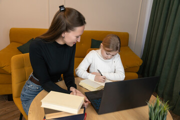 Mother and daughter are studying together at home, with the mother helping the daughter with her homework while the child writes in a notebook, creating a supportive learning environment
