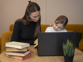 Mother and daughter studying together, focusing on a laptop screen, engaging in online education for school homework while using a notebook for learning and tutoring at home
