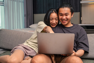 Asian couple sitting on a sofa smiling while looking at a laptop together, appearing happy and relaxed in a bright bedroom with natural daylight through the window