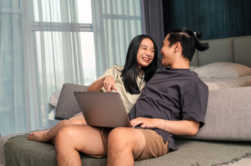 Asian couple laughing joyfully together while sitting on a couch with a laptop, showing affection and happiness in a cozy bedroom with daylight coming through the window