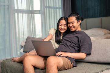 Asian couple sitting on a sofa smiling while looking at a laptop together, appearing happy and relaxed in a bright bedroom with natural daylight through the window