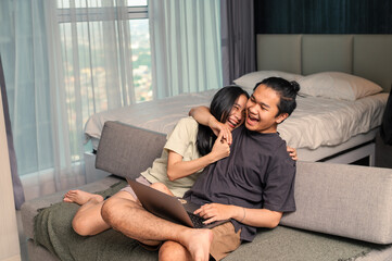 Asian couple laughing joyfully together while sitting on a couch with a laptop, showing affection and happiness in a cozy bedroom with daylight coming through the window