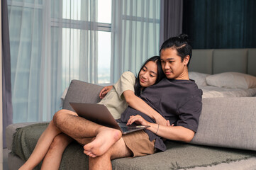 Obraz premium Asian couple sitting closely on a couch, cuddling while watching something on a laptop, sharing a calm and affectionate moment together in a cozy modern apartment with daylight