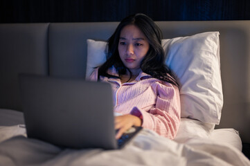 Asian woman in pink pajamas lying on bed and using a laptop with a focused expression, illuminated by warm bedroom light at night