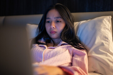 Asian woman in pink pajamas lying on bed and using a laptop with a focused expression, illuminated by warm bedroom light at night