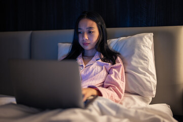 Asian woman in pink pajamas lying on bed and using a laptop with a focused expression, illuminated by warm bedroom light at night