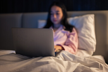 Asian woman in pink pajamas lying on bed and using a laptop with a focused expression, illuminated by warm bedroom light at night