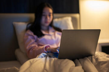 Asian woman in pink pajamas lying on bed and using a laptop with a focused expression, illuminated by warm bedroom light at night