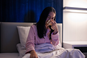 Asian woman in pink pajamas sneezing into a tissue while sitting on bed, looking unwell with signs of cold or allergy in a dim bedroom