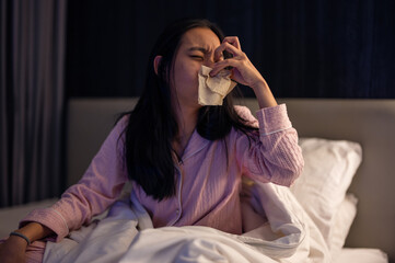 Asian woman in pink pajamas sneezing into a tissue while sitting on bed, looking unwell with signs of cold or allergy in a dim bedroom