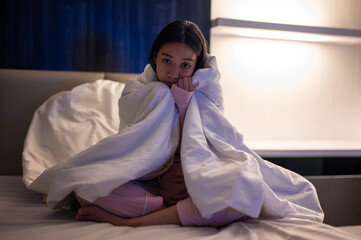 Asian woman wrapped tightly in a white blanket, sitting on bed with eyes closed, appearing cold or unwell in a dimly lit bedroom at night