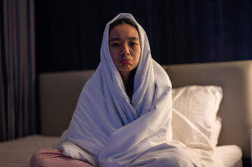Asian woman wrapped in white blanket sitting on bed with pouty face, looking sad and uncomfortable in dimly lit bedroom