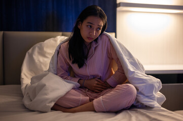 Asian woman sitting on bed wearing pink pajamas and covered with a white blanket, holding her stomach and looking unwell in a dimly lit bedroom