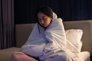 An Asian woman wrapped tightly in a white blanket, sitting on a bed with eyes closed and a pained expression, looking cold, unwell, or emotionally distressed