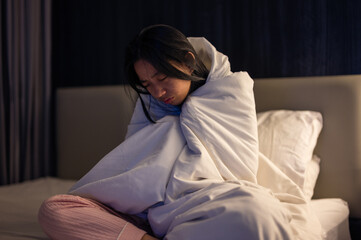 An Asian woman wrapped tightly in a white blanket, sitting on a bed with eyes closed and a pained expression, looking cold, unwell, or emotionally distressed