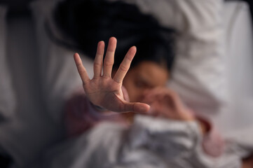 An Asian woman lying in bed with white pillows and blanket, wearing pink pajamas, extending her hand forward as if to block the camera, showing tiredness, stress, or refusal to be disturbed