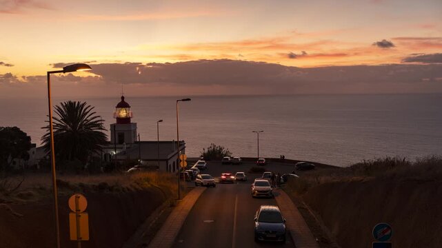 Wide timelapse view of Farol da Ponta do Pargo lighthouse at dusk on Madeira Island, Portugal. The access road and parking area lead toward the coastal lighthouse as its rotating light beam sweeps sky