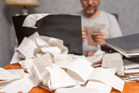 Overwhelmed man reviewing receipts behind a desk covered with scattered papers and folders. Concept of financial stress, expense tracking, paperwork overload, and budgeting problems.