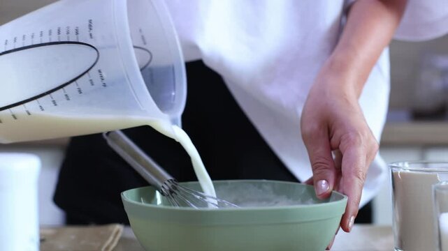 A person pours milk from a measuring jug into a green bowl of dry ingredients, then whisks it together to form a smooth, creamy batter for baking.