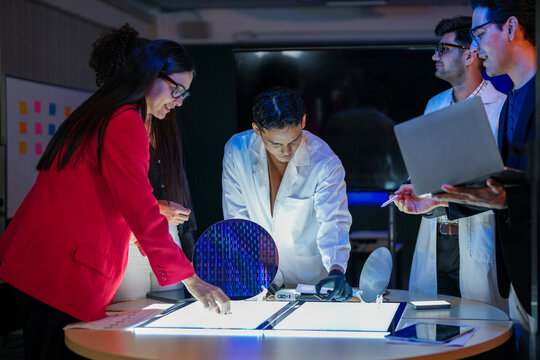 Specialist team of engineers and executives inspecting a large silicon wafer in a high-tech semiconductor laboratory. Discussion on microchip and RAM memory manufacturing process and quality control. - Powered by Adobe