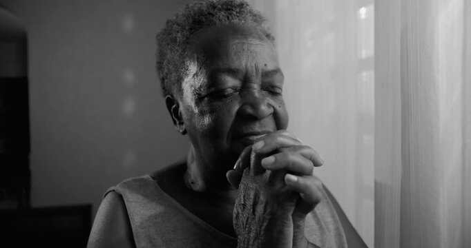 Elderly African American woman praying by window at home, quiet faith, seeking comfort, spiritual reflection, calm perseverance in later life