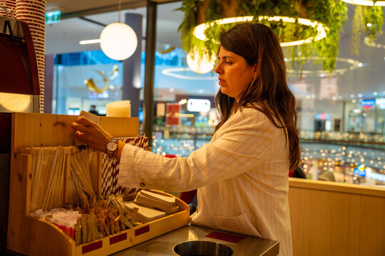 Woman selecting napkins, straws and disposable cutlery at cafe self service bar after ordering. Food service routine, consumer choice and modern lifestyle moment in real life commercial environment