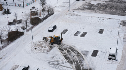 Heavy machinery clearing snow from a parking lot, showcasing winter weather challenges, with vehicles and trees in the background, emphasizing the importance of snow removal