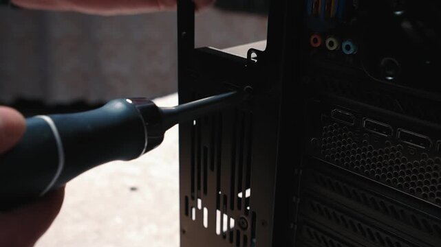 Unscrewing the hard drive and SSD bracket mount from the back panel of the PC case, close-up of screwdriver and screw next to I/O ports
