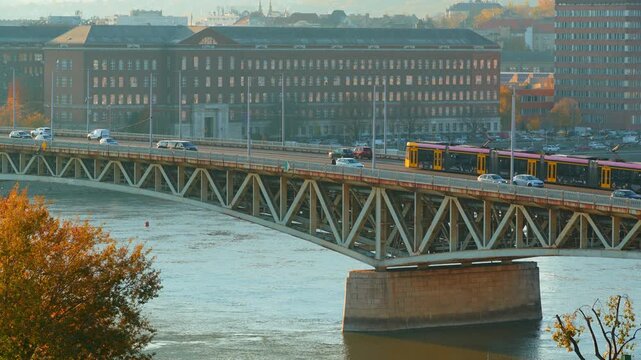 Panoramic aerial view of a busy Budapest street in autumn, featuring architecture and Petofi bridge, yellow fall trees, city traffic with cars and buses, and pedestrians walking on sunny sidewalks