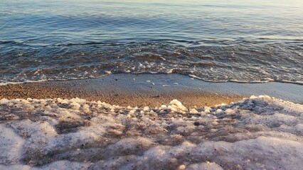 Snowy Shoreline and Freezing Sea. A gentle wave rolls onto a snowy beach, creating a serene contrast between water and snow. The sunset casts a warm light over the scene.