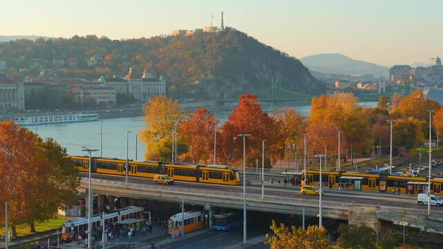 Panoramic aerial view of a busy Budapest street in autumn, featuring architecture and Petofi bridge, yellow fall trees, city traffic with cars and buses, and pedestrians walking on sunny sidewalks
