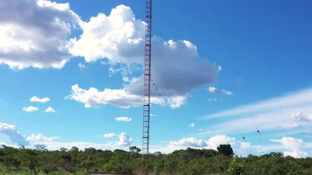 Vertical antenna mast with diagonal bracing and warning stripes rises against cumulus cloud backdrop with dense green cerrado forest surrounding base elevation.
