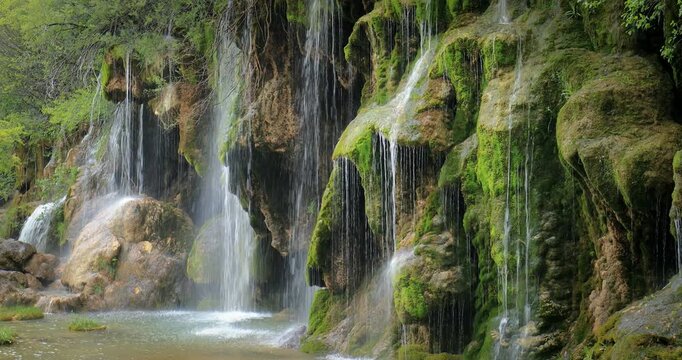 Cinematic view of waterfall flowing gently over moss covered rocky cliff with thin streams and soft mist creating serene mood, rich texture, and peaceful natural scenery