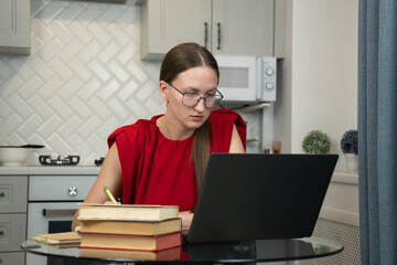 Young woman wearing glasses and a red shirt, taking notes while studying at home. Working on a...