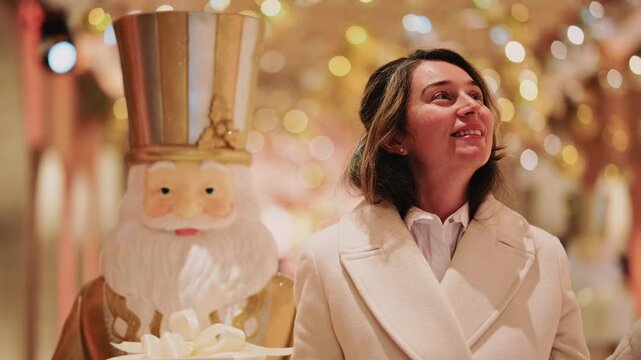 A woman in a light colored coat stands beside a large nutcracker decoration, surrounded by warm festive lights