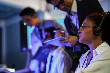 Cyber security analysts monitoring network threats in a blue-lit control room. IT specialists with headsets discuss digital safety and system defense data on a tablet device.