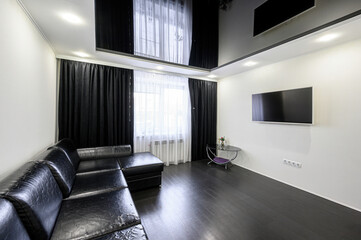 Fototapeta premium Modern living room with black leather sectional sofa, wall-mounted TV, dark wooden floor, black curtains, sheer white drapes, small glass side table with decor