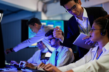 Cyber security analysts monitoring network threats in a blue-lit control room. IT specialists with headsets discuss digital safety and system defense data on a tablet device.
