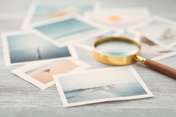 Brass magnifying glass with wooden handle on pile of scattered instant print photos depicting various beach and ocean scenes,on light wooden table