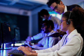 Cyber security analysts monitoring network threats in a blue-lit control room. IT specialists with headsets discuss digital safety and system defense data on a tablet device.