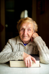 Portrait of an elderly woman resting her hand on a book indoors, looking at the camera with a calm expression, soft natural light highlighting her face and knitted sweater.