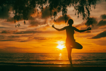 Silhouette of a person standing in a yoga pose on a beach at sunset, with the sun low over the sea, dramatic clouds, and tree branches framing the scene.