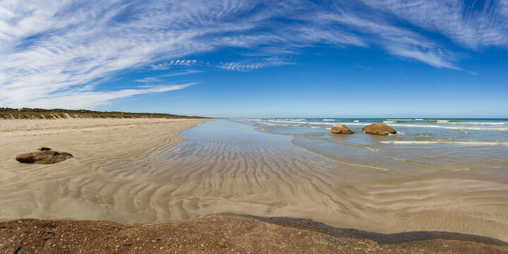 Aerial panorama view of waves lapping against large granite boulder embedded in a rippled sand beach
