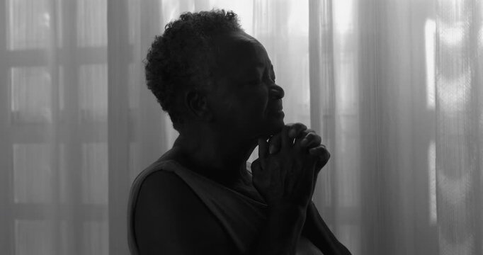 Elderly African American woman praying softly by window in monochrome, eyes closed, enduring faith, quiet hope, spiritual perseverance in later life