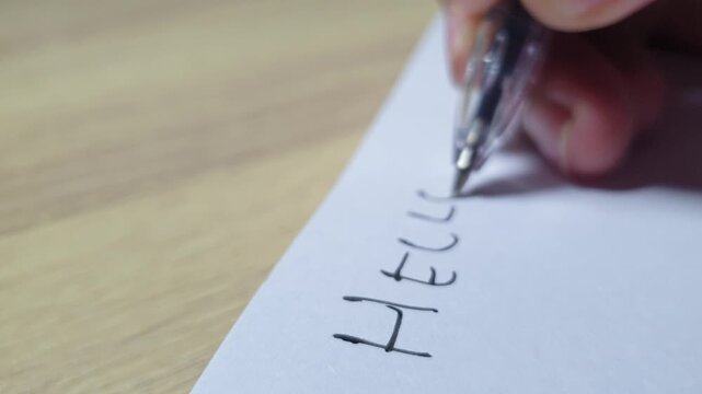 Close up footage of a hand writing the word Hello on white paper with a black pen, illustrating communication, greeting, welcoming concepts, and starting a letter.