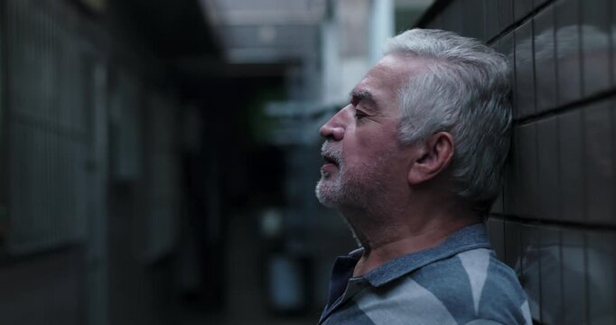 Senior man leaning against wall in narrow corridor with eyes closed, exhausted pause, emotional weight, quiet endurance during difficult times