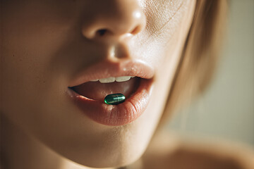 Close-up of glossy lips holding a translucent green capsule between teeth, conveying medication, supplements and sensual beauty lifestyle